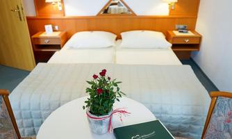 A cozy hotel room with two single beds and warm lighting. On the table, there is a pot of red roses and a welcome booklet.