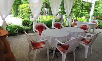 An inviting garden area with a round table and white plastic chairs. The surroundings are green and well-planted, ideal for a relaxing time outdoors.
