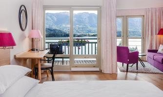 A modern room with a view of the mountains and a lake. The decor is stylish with a desk, a cozy chair, and vibrant colors.