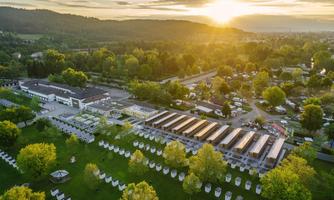 A picturesque landscape with a campsite and modern accommodations. The sun rises over the hills and illuminates the green surroundings.