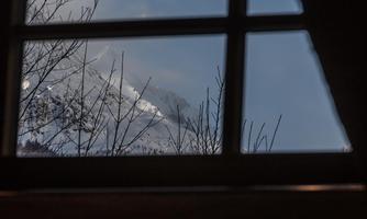A view through a window of snow-covered mountains and a cloudy sky. Branches of a tree are visible in the foreground.