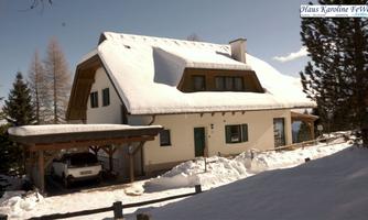 A snow-covered house in alpine style with a separate carport. Surrounded by fir trees and a serene winter landscape.
