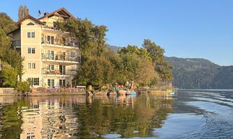 A picturesque house by the shore of a crystal clear lake. The surroundings are green and tranquil, with gentle hills visible in the background.