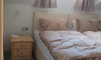 A cozy bedroom with a large bed and a small nightstand. The walls are bright, and the light falls through the window.
