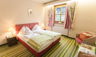 A cozy hotel room with a double bed, two bedside tables, and an armchair. Bright curtains and a large window front create a friendly atmosphere.