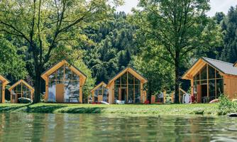 Modern wooden cabins by the shore of a calm water. Surrounded by green trees and a picturesque landscape.