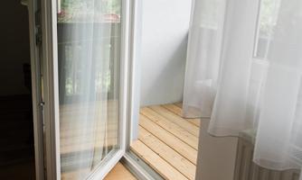 An open window with a balcony view. The floor is made of light wood and the curtains are slightly transparent.