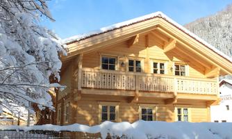 A cozy wooden house in the snow with a balcony. The surroundings are surrounded by snow-covered trees.