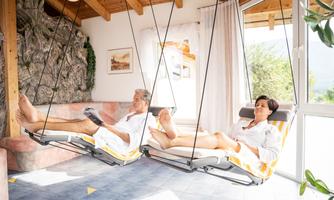 A couple relaxes in a modern hammock in a bright room. The atmosphere is calm and inviting, with plenty of natural light.