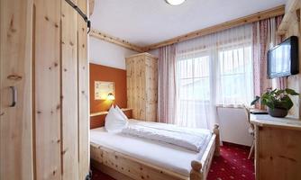 A cozy room with wooden furniture and a single bed. Large windows let in plenty of light and there is a TV and plant decoration.