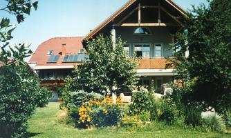 A modern house surrounded by a well-maintained garden with colorful flowers and trees. The sun shines on the roof with solar panels.