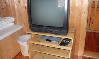 An old television on a wooden table in a simple room. In the background, bed linen and a wastebasket can be seen.