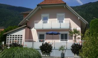 A pretty pink house with a red roof and a balcony. The garden is well-kept with plants and a sun umbrella.