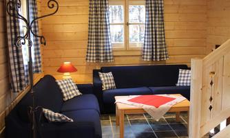 A cozy living room with wooden walls and a blue sofa. The room has large windows and a natural stone floor.