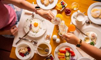 A festive breakfast with a variety of foods and drinks on a table. Two people clink their glasses and enjoy the meal together.