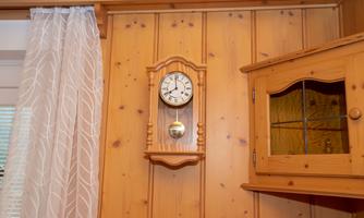 A wooden wall clock hangs on a wooden paneling. Next to the clock is a cabinet with a glass front.
