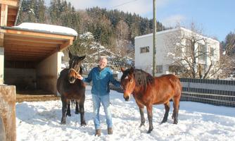 A man stands in the snow holding two horses on a leash. In the background, a house and trees can be seen.