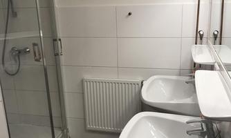 A modern bathroom with two sinks, a shower, and a radiator. The walls are tiled in white and the floor is in shades of gray.