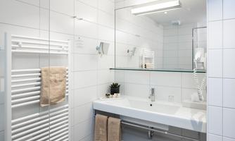A modern bathroom with white tiles and a large mirror. It includes a sink, towels, and a towel rack.