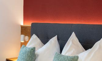 A cozy bedroom with a dark blue headboard and white pillows. The wall is painted in a warm shade of red and there are decorative cushions in soft colors.