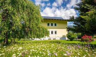 A beautiful house surrounded by colorful flowers and green trees. The sky is clear and the sun is shining.