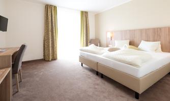 A modern hotel room with two beds and a desk. Large windows provide plenty of daylight.
