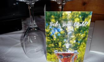 An elegant table with two inverted wine glasses and a card that says "Cheers!" In the background, blurred green trees can be seen.