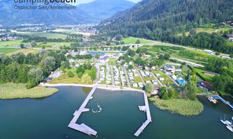 An aerial view of Lake Ossiach with campsites and a small harbor. Surrounded by green forests and gentle hills, the landscape offers a peaceful atmosphere.