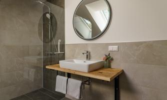 A modern bathroom with a large round mirror and an elegant wooden washbasin console. The shower is separated by glass, and neutral tones create a calming atmosphere.