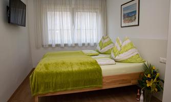 A cozy bedroom with a double bed and green pillows. The windows are covered with light curtains, and a splash of color comes from a small plant.