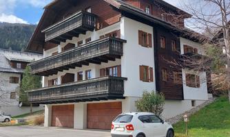 A modern chalet with wooden cladding and balconies. In front of it is a car, surrounded by nature and mountains.