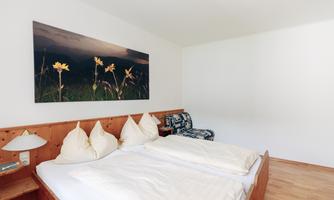 A cozy bedroom with a double bed and bright pillows. On the wall hangs a picture with yellow flowers.