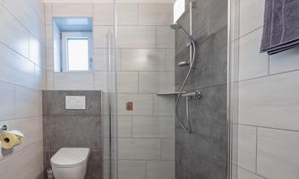A modern bathroom with a shower and a toilet. The walls are tiled in light colors, and a window provides daylight.