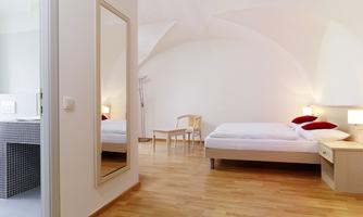 A bright bedroom with two beds and red pillows. The floor is wooden and a mirror is leaning against the wall.