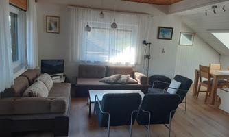 A cozy living room with a couch and armchairs. There is a dining table and plenty of daylight through the windows.
