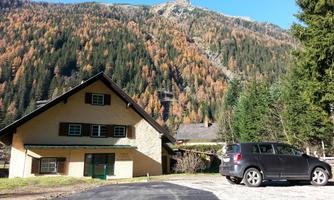 A charming house in the mountains, surrounded by colorful trees in autumn. A black car is parked on the paved path.