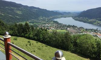 A picturesque view of a lake surrounded by hills and trees. In the background, small houses and a gentle landscape can be seen.