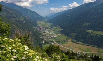 A picturesque mountain landscape with green valleys and a small village. White clouds drift across the clear blue sky.