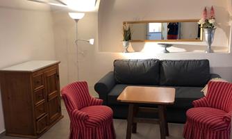 A cozy living area with a gray sofa, a wooden table, and two red chairs. There is a dresser against the wall and decorative flowers on the table.