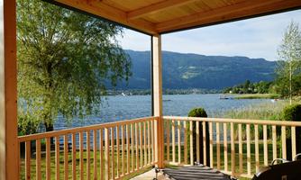 A wooden porch overlooking a tranquil lake and the surrounding mountains. The scene is surrounded by trees and green grass, conveying a peaceful atmosphere.