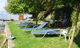 A quiet garden with loungers on the lawn and a view of the water. In the background, trees and a seating area can be seen.