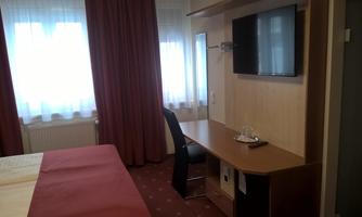 A cozy hotel room with a desk, a TV, and large windows. The walls are decorated with red fabric, and the floor is carpeted.