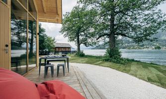 A modern house by the water with a terrace and a dining table. In the background, there are trees and the serene landscape.