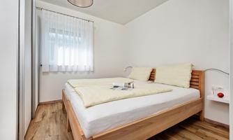 A cozy bedroom with a wooden bed and two pillows. The room has light walls, a modern lamp, and a large window with curtains.