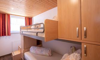 A cozy room with a wooden bunk bed and a single bed. Large windows provide plenty of daylight.