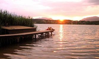 A tranquil lake with a wooden jetty and green reeds. The sun sets on the horizon, creating a picturesque atmosphere.