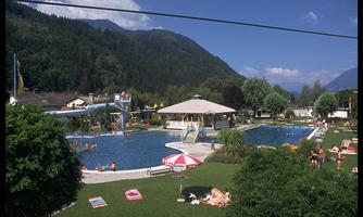 A large pool area with a slide and several swimming pools. Surrounded by green meadows and mountains, people enjoy the sunny day.