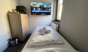 A simple bedroom with a single bed and a television on the wall. Next to the bed is a small wardrobe and a trash can.