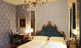 An elegant bedroom with an artistically adorned bed and a bright color palette. The wall is decorated with a classic pattern, and a chandelier creates a stylish atmosphere.