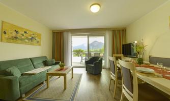 A bright and modern living room with a green couch and a view of the mountains through the windows. The space is stylishly furnished with a dining table and decorative elements.